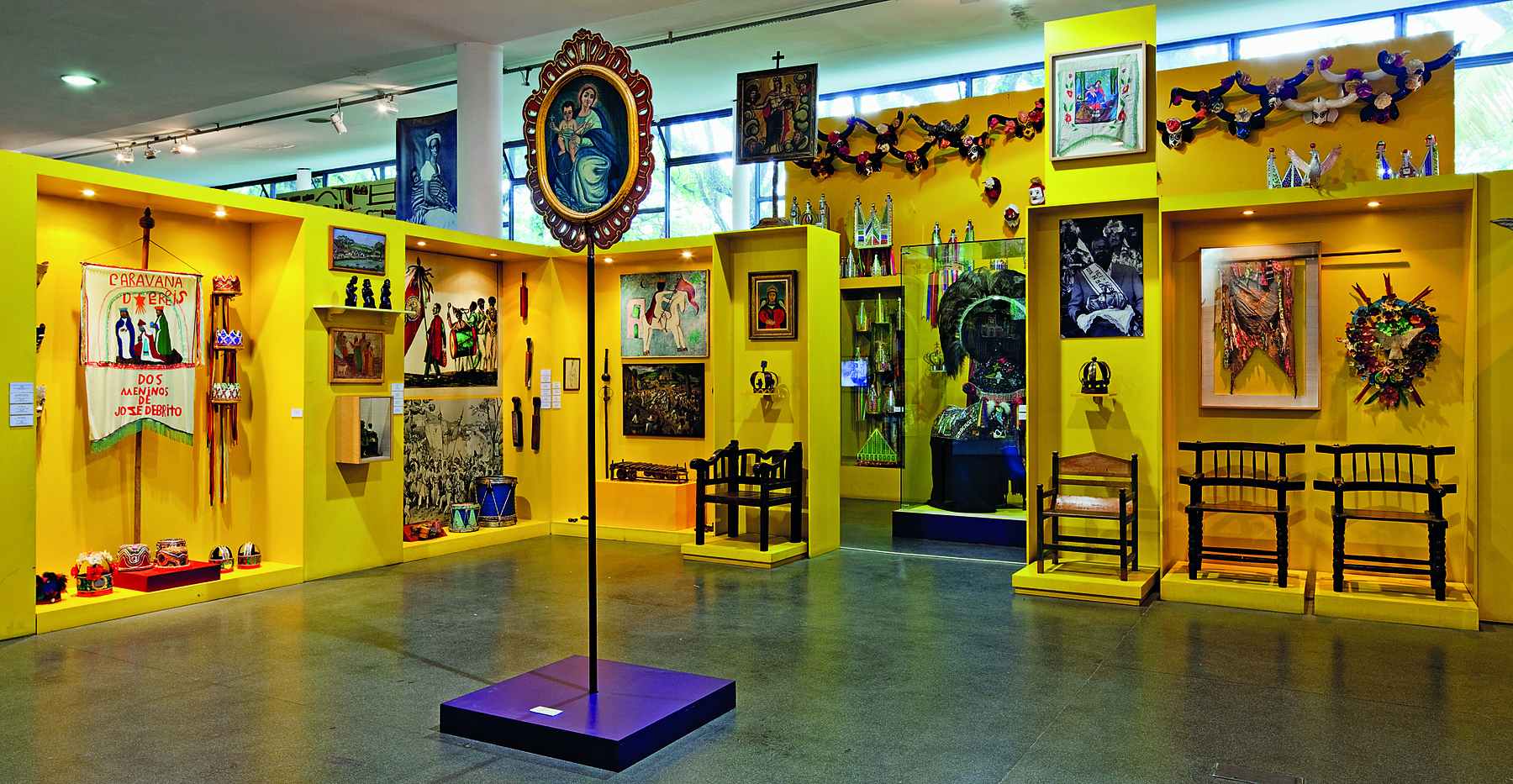 Fotografia. Uma das salas do Museu Afro Brasil. Em primeiro plano, um retrato com moldura arredondada e dourada suspenso em um pedestal com haste fina. Em segundo plano, prateleiras amarelas, com adereços e objetos pendurados.