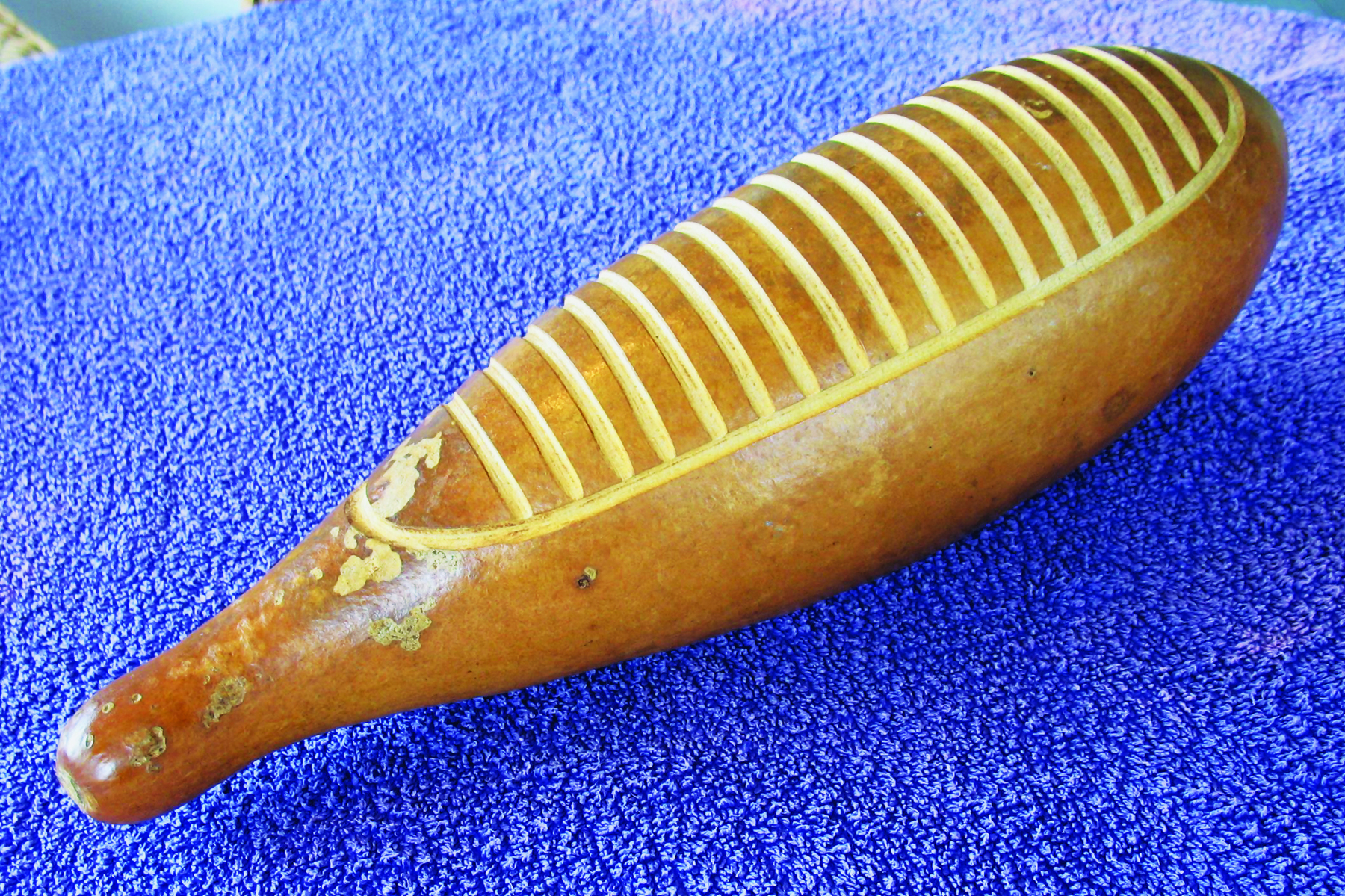 Fotografia. Um instrumento musical de percussão feito de madeira. Ele tem formato horizontal e arredondado, com uma das pontas mais finas. Acima, algumas linhas em branco.