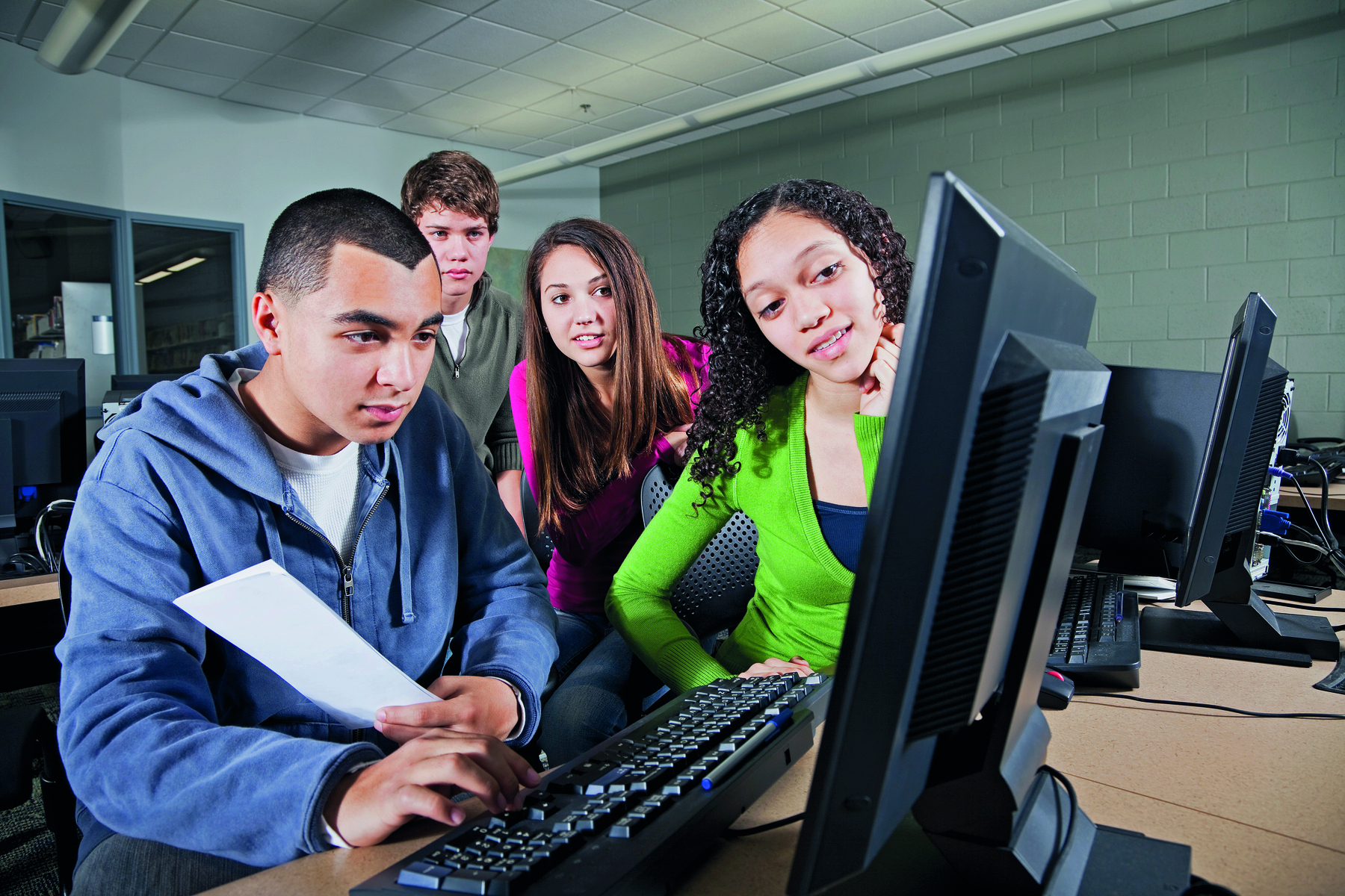 Fotografia. Um grupo de quatro jovens estão juntos e olhando para a tela de um computador. Um deles está mais à frente e digita no teclado, enquanto segura uma folha em uma das mãos. Ao lado dele, os demais olham atentamente. Eles vestem roupas azuis, verdes, rosa e cinza.