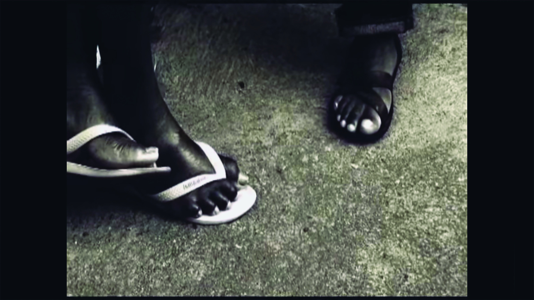 Cena de filme. Cena em preto e branco. Vista do alto de pés de duas pessoas pisando em um chão de concreto cinza. À esquerda, dois pés cruzados, usando chinelos claros e, à direita, um pé usando sandália escura.
