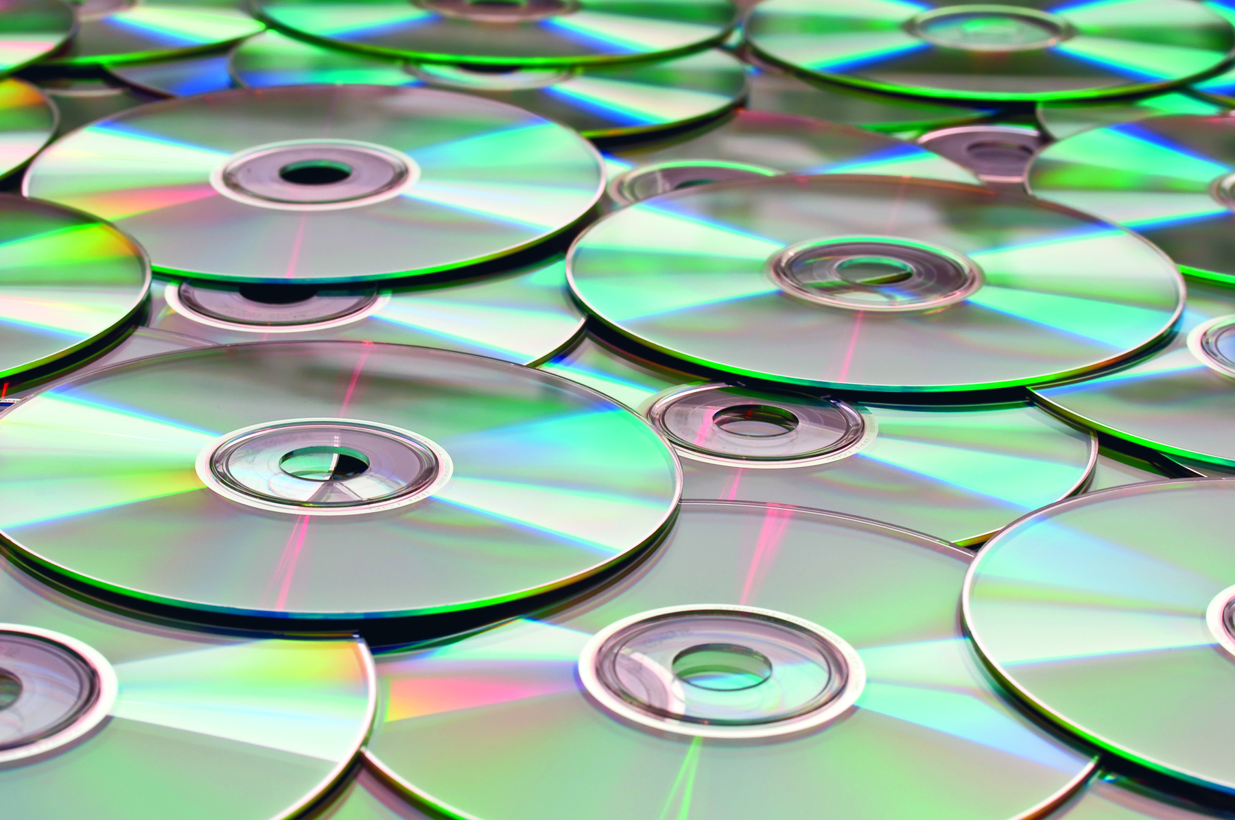 Fotografia. Foco em CDs ou DVDs espalhados um sobre outro, de formato redondo em tons de cinza e reflexos em verde-claro.