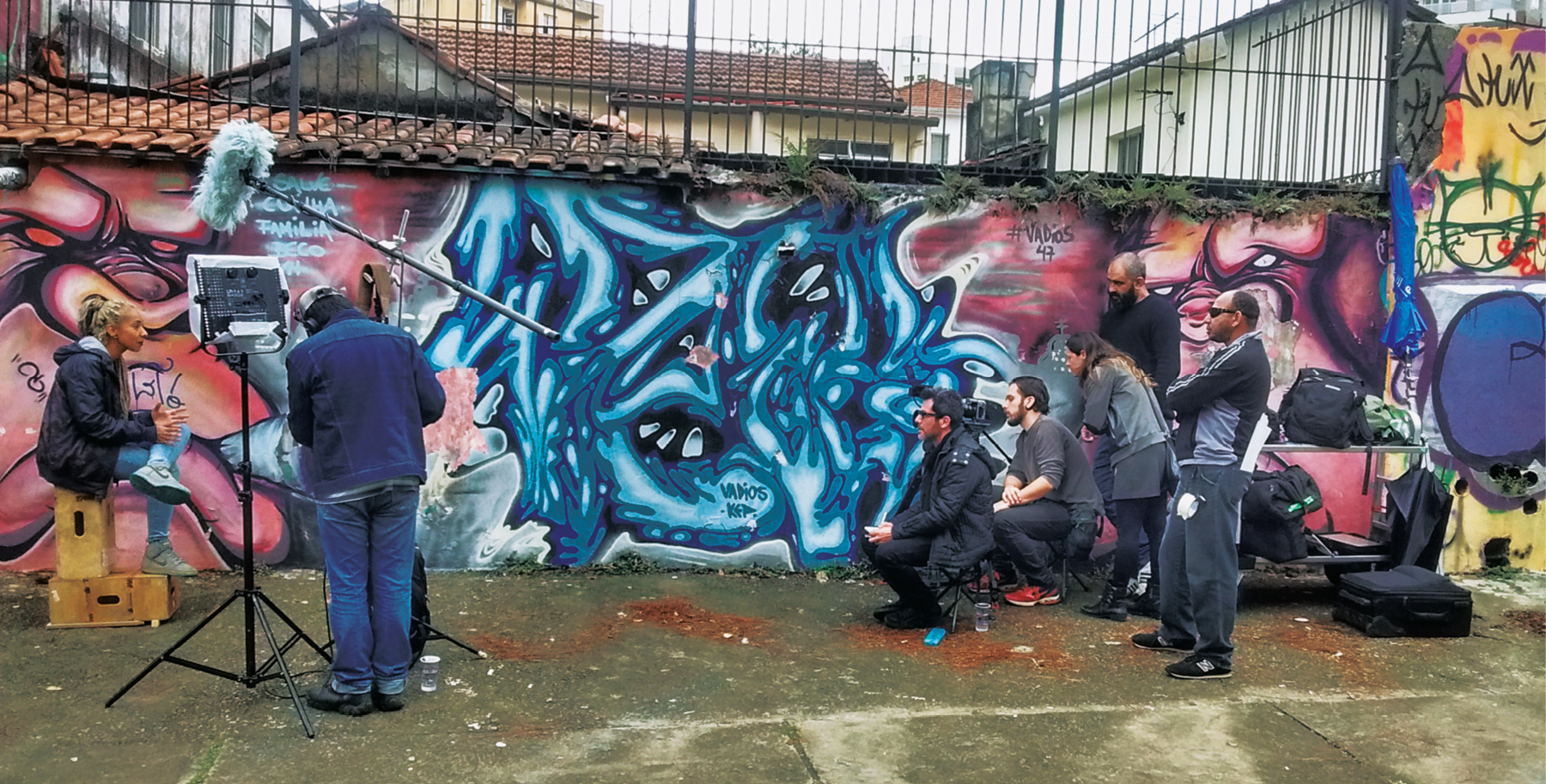 Fotografia. Realização de  filmagem de documentário em espaço externo. À esquerda, uma mulher sentada sobre caixotes de madeira; ela veste calça jeans, casaco preto e tênis. Perto dela, um homem visto de costas, de jaqueta, calça jeans e sapato preto; ele está diante de um tripé com pontos de iluminação e microfones no alto, na direção da mulher. À direita, dois homens agachados, de camiseta e calça preta e outras três pessoas em pé, também com roupas escuras. Todos observam a mulher na outra ponta. Um dos homens está com uma filmadora. Ao fundo, um muro com grafite abstrato em tons de rosa, azul e preto.