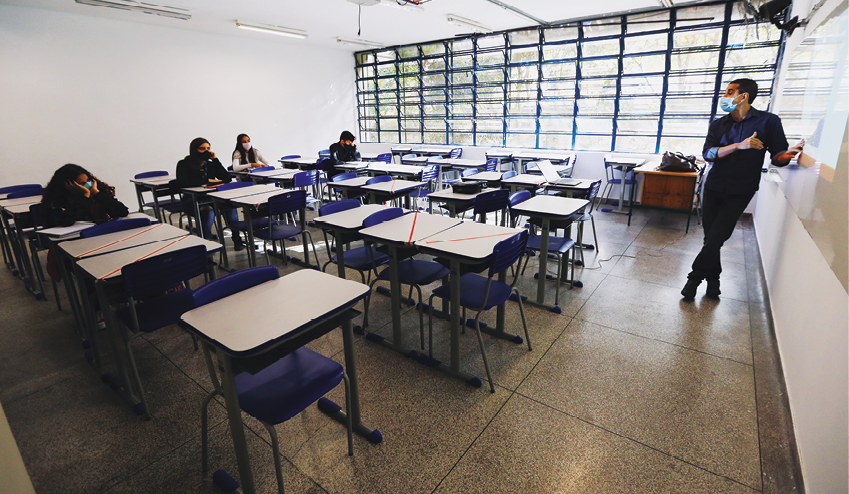 Fotografia. Vista lateral de uma sala de aula. Em destaque, um professor diante da lousa, à direita, e diversas mesas bege com cadeiras azuis à esquerda. Alguns alunos estão sentados nas cadeiras do fundo, mas muitos assentos estão vagos. Ao fundo, uma grande janela de vidro cobrindo a parede toda.