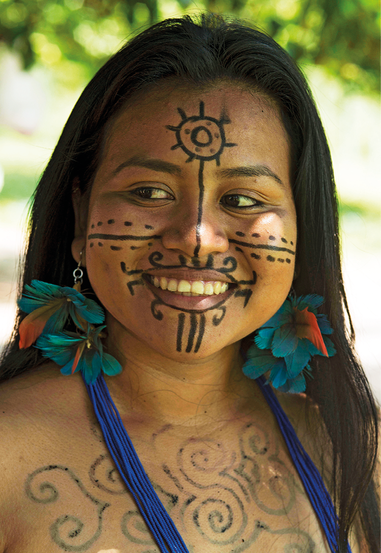 Fotografia. Destaque de uma mulher indígena com o rosto pintado com linhas pretas: na bochecha, há linhas horizontais retas e tracejadas; ao redor da boca e na testa, linhas circulares; no queixo, linhas retas na vertical.