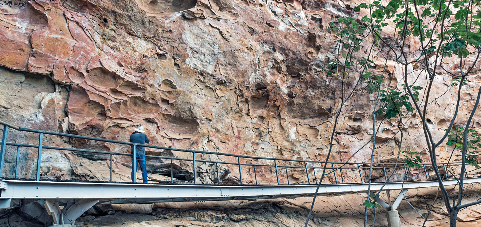 Fotografia. Destaque para uma ponte margeando uma montanha marrom com diversas pedras. No centro da ponte, há uma pessoa em pé.