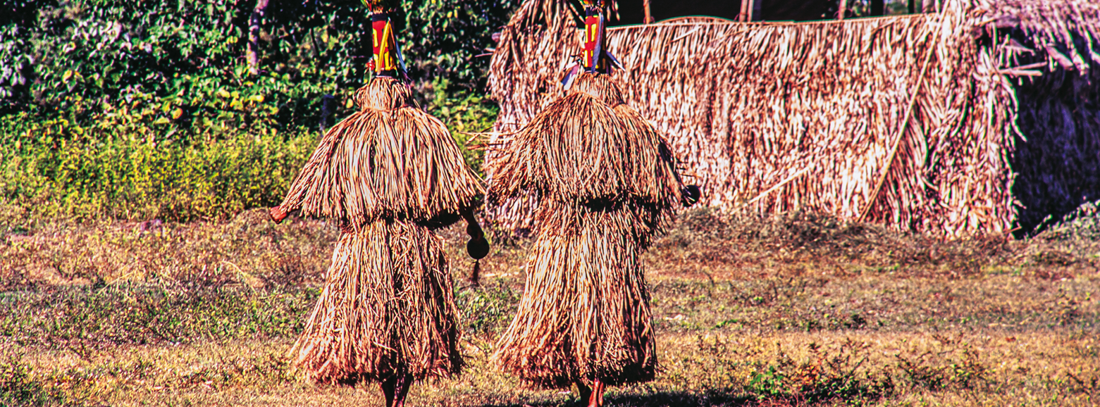 Fotografia. Destaque para duas pessoas ao centro da imagem usando roupas de feno cobrindo o corpo inteiro e a cabeça. Elas estão correndo na grama na direção de uma casa retangular de bambu e feno. Ao fundo, uma vegetação verde.