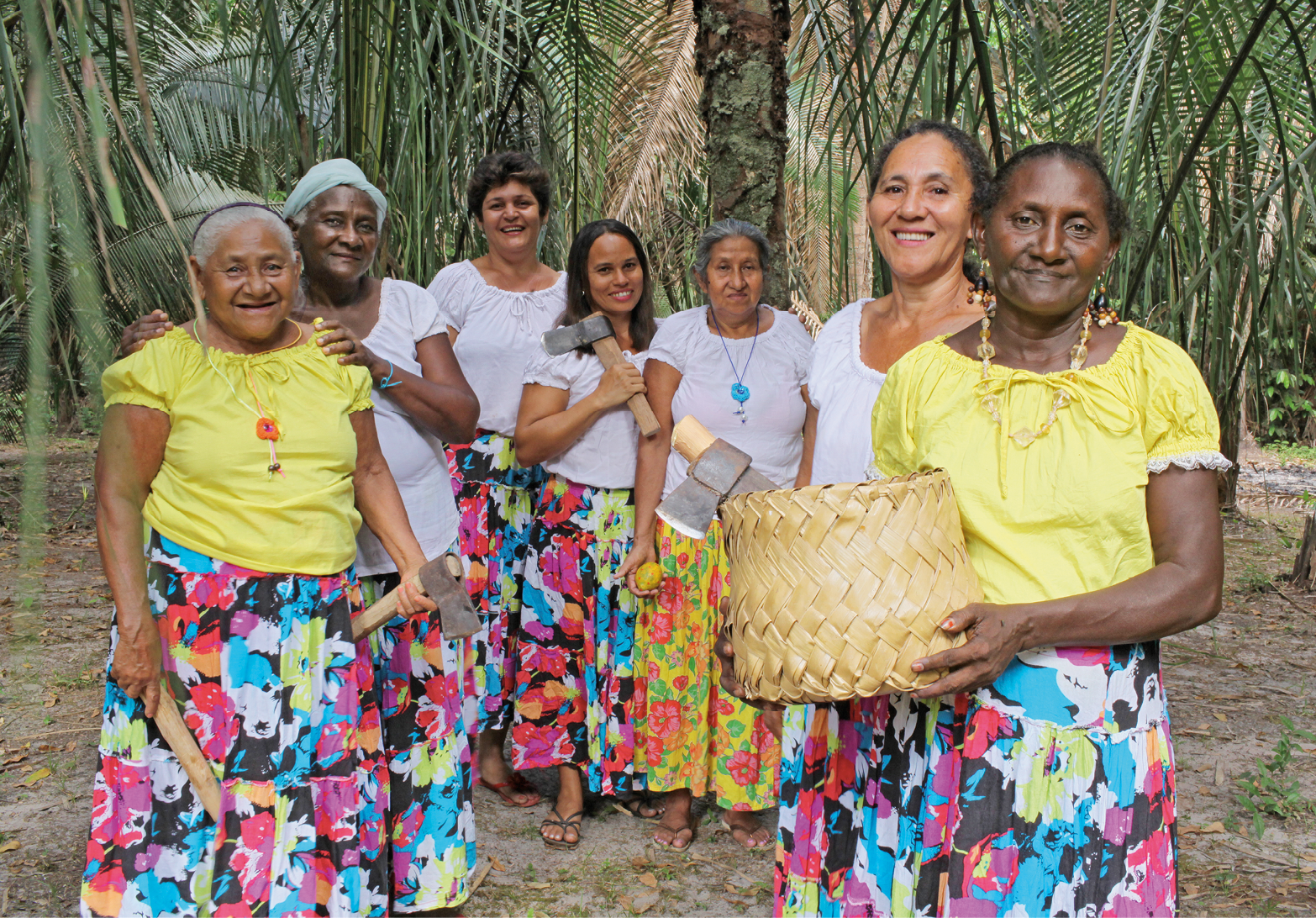 Fotografia. Sete mulheres em pé, usando camisetas brancas ou amarelas e saias coloridas; algumas seguram machados pequenos; a que aparece em primeiro plano segura um cesto redondo e bege. Ao fundo, há árvores verdes.