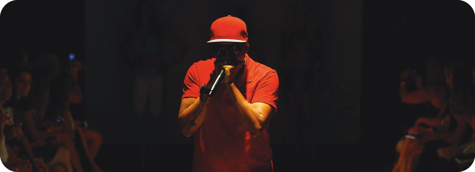 Fotografia. Um homem de boné e camisa vermelha, segurando um microfone na frente da boca, está com partes do corpo iluminadas e o rosto no escuro; ao fundo, há uma plateia na escuridão.