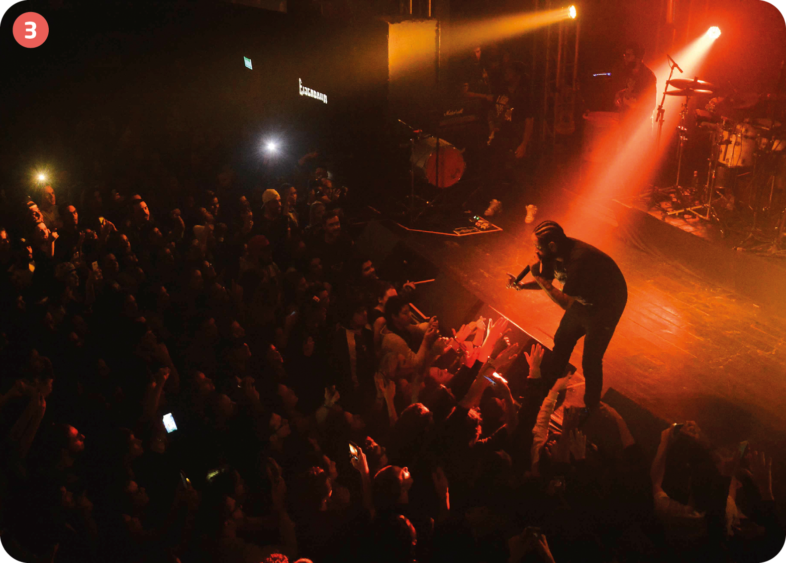 Fotografia. Vista lateral de cima de um palco iluminado com luzes vermelhas; à direita, há um homem com cabelo penteado com tranças para trás e segurando um microfone na frente da boca. À esquerda, há uma plateia no escuro.