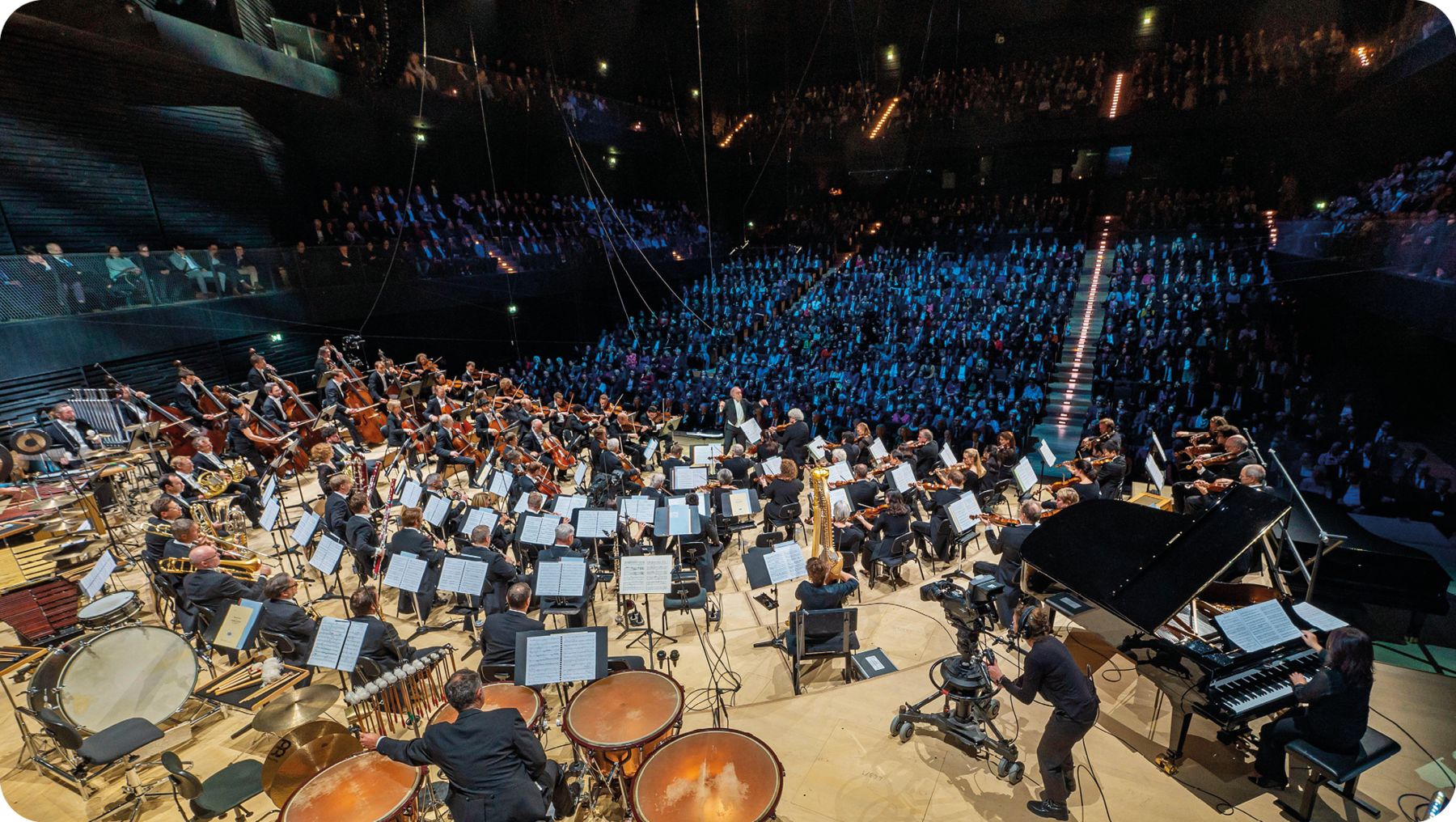 Fotografia. Vista de cima de uma orquestra iluminada e com diversos músicos sentados e tocando diferentes instrumentos musicais. No canto inferior direito, há um piano preto. Ao fundo, uma plateia assiste ao concerto.