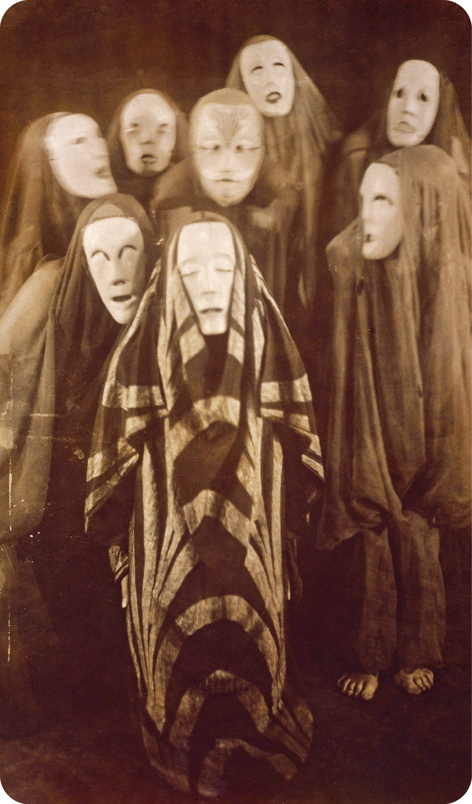 Fotografia em preto e branco. Sete pessoas em pé e vestidas com fantasias de pano preto longo e uma máscara branca com olhos fechados.