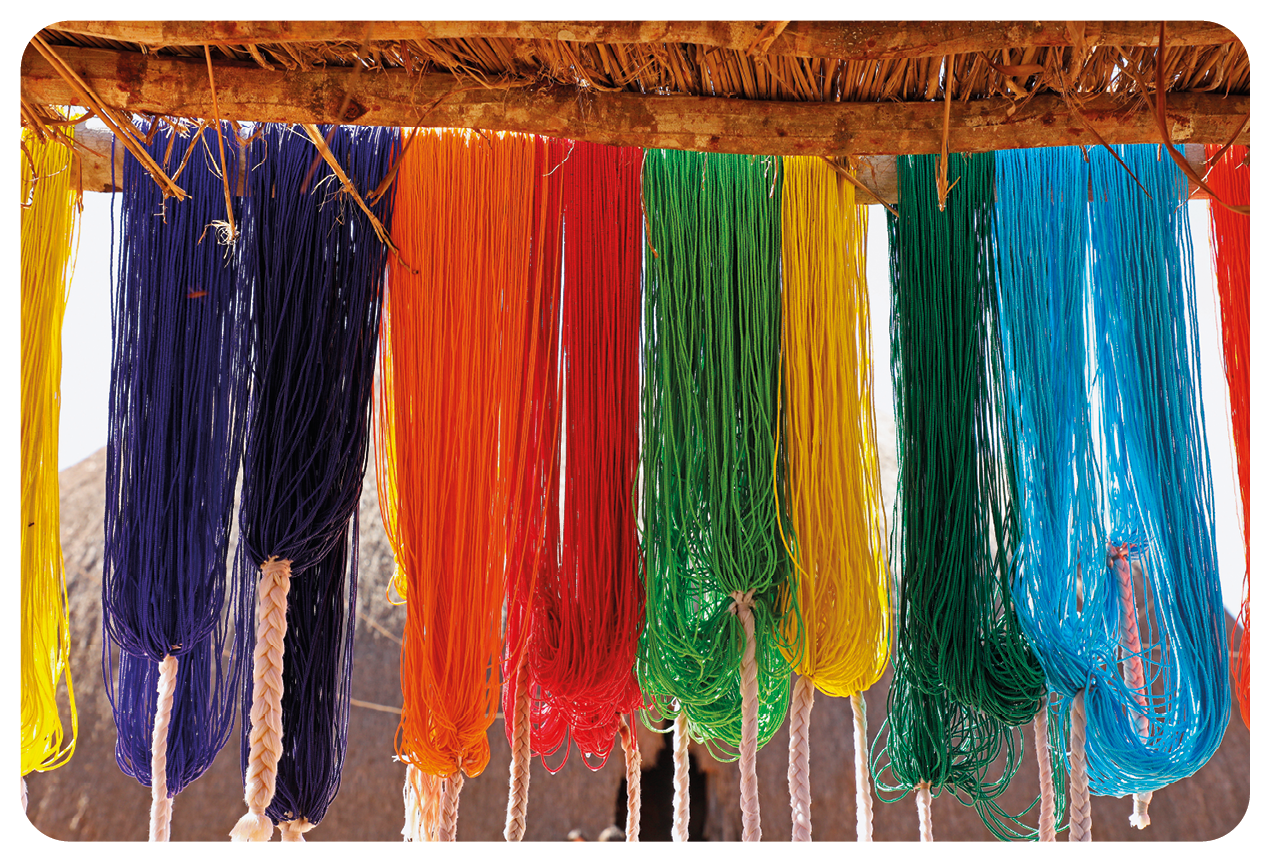 Fotografia. Destaque para um varal com diversos adereços de fios coloridos em tons de roxo, laranja, vermelho, verde, amarelo e azul.