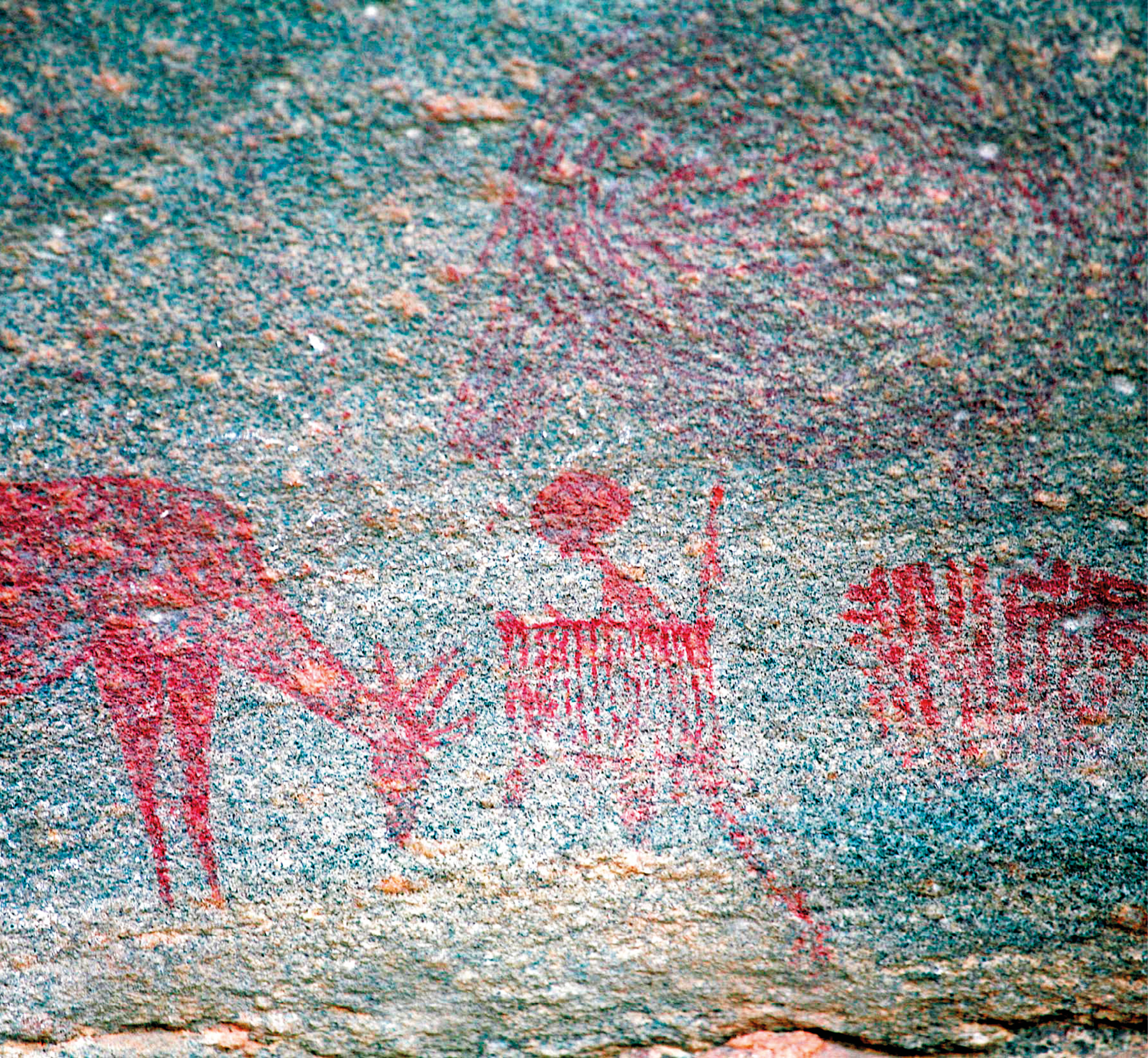 Fotografia. Destaque para uma parede de caverna em tons de azul; sobre ela há uma pintura rupestre em tons de vermelho retratando formas humanas e animais.