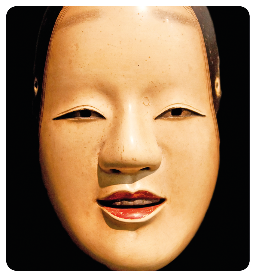 Fotografia. Destaque para uma máscara de madeira com olhos finos, nariz largo e lábios finos.
