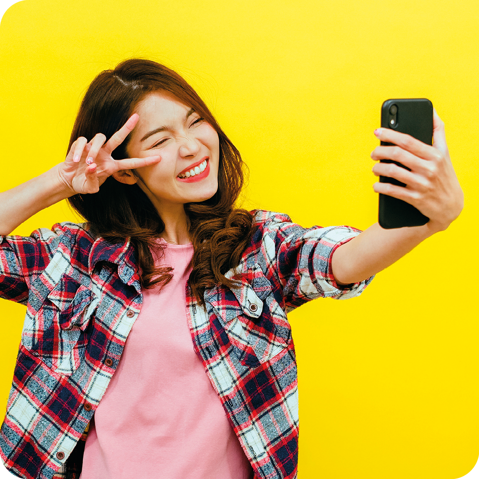 Fotografia. Uma menina de cabelo castanho penteado para o lado, de olhos puxados, nariz e lábios finos, usando blusa xadrez aberta e camiseta rosa, está tirando selfie com o celular, fazendo pose e sorrindo.