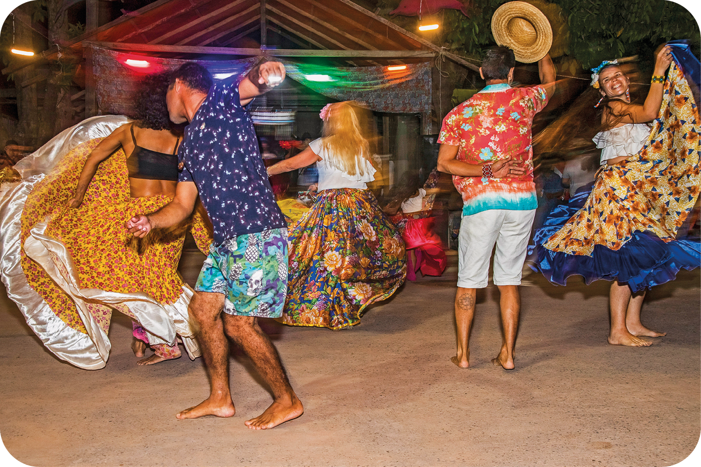 Fotografia. Dois homens usando camisa e shorts e três mulheres usando camisa e saia comprida colorida estão dançando. As mulheres movimentam as saias, um dos homens tira o chapéu e uma delas sorri.