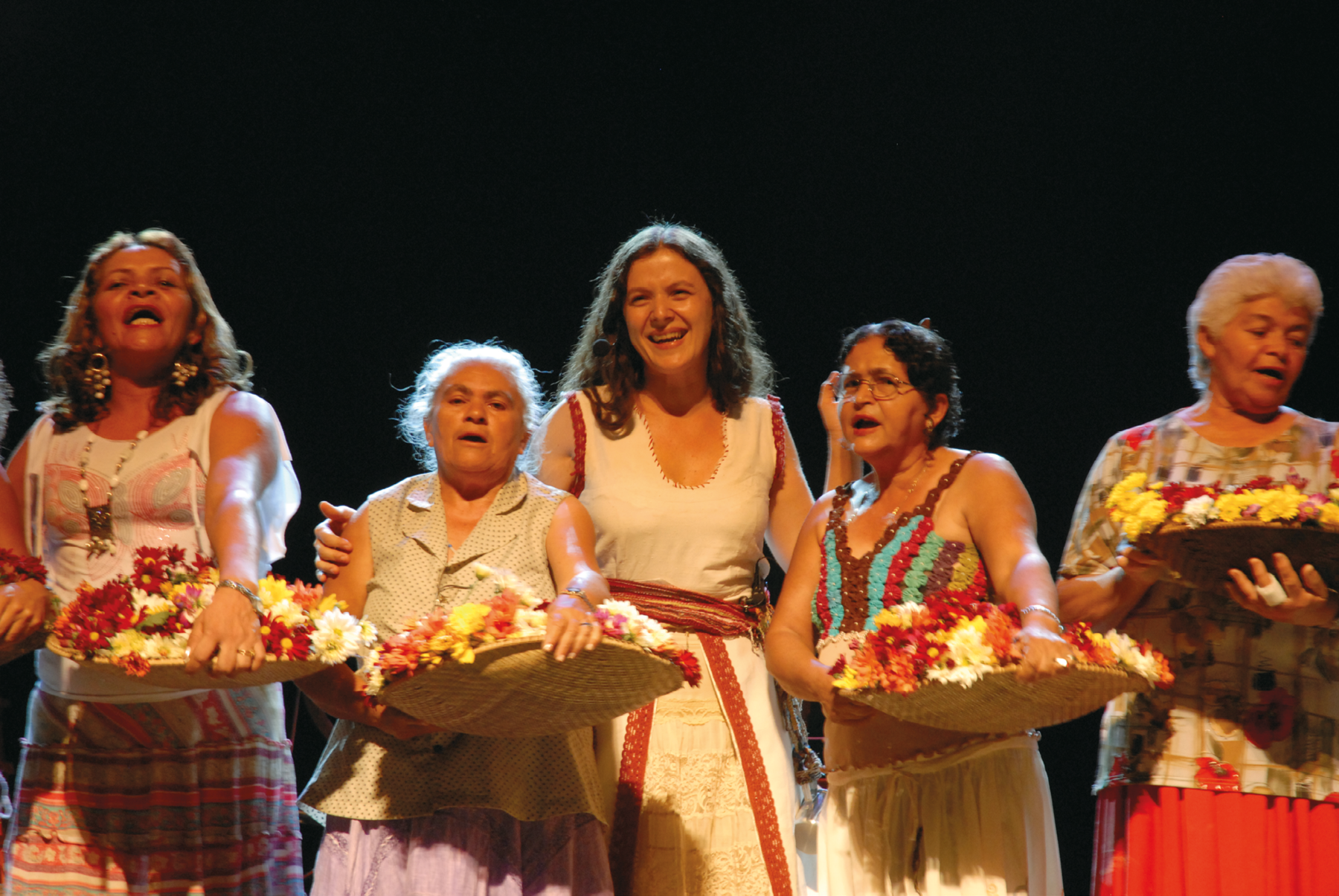 Fotografia. Cinco mulheres perfiladas e sorrindo; todas estão em pé e quatro delas seguram um cesto redondo cheio de flores. Ao centro, há uma mulher usando vestido branco e sorrindo, enquanto as demais cantam.