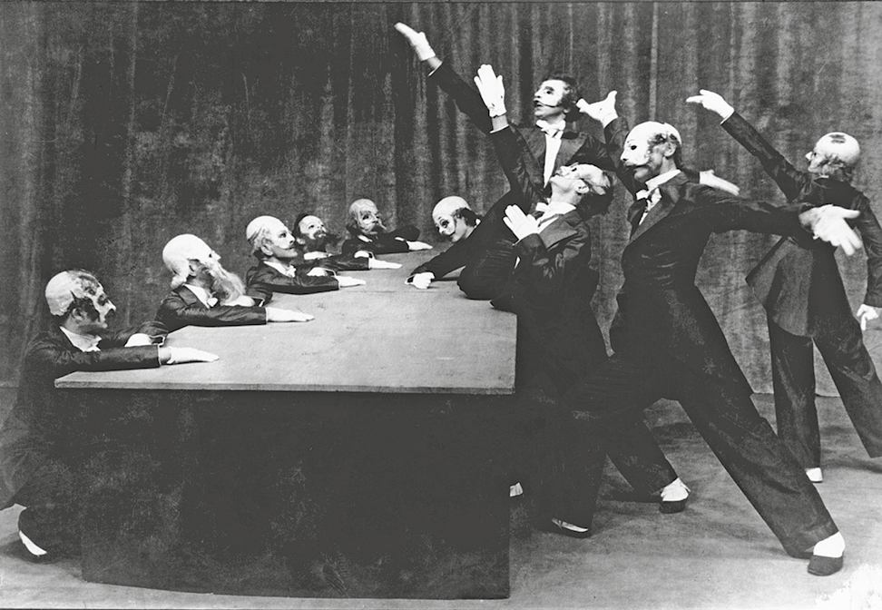 Fotografia em preto e branco. À esquerda, cinco homens usando roupa social estão sentados e com as mãos apoiadas sobre uma mesa larga e preta; à direita, diversos homens usando terno preto estão em pé, inclinados na direção da mesa e com os braços levantados.