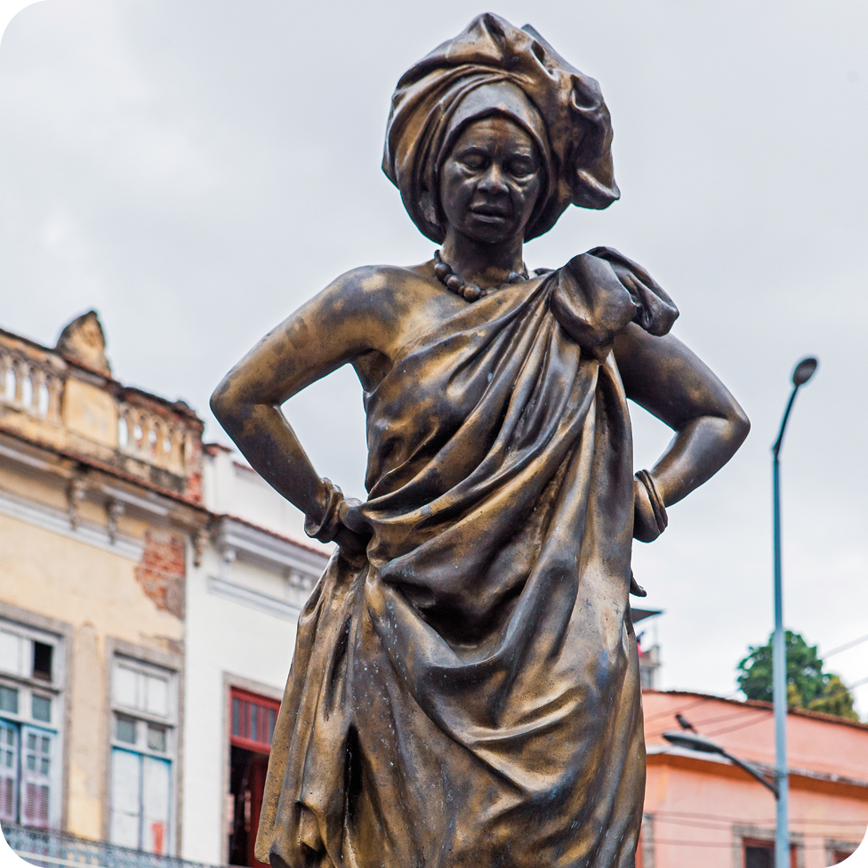 Fotografia. Destaque para uma estátua em tons de bronze escuro; uma mulher de nariz largo, usando lenço dobrado na cabeça e um vestido amarrado no ombro; ela está com as duas mãos na cintura.