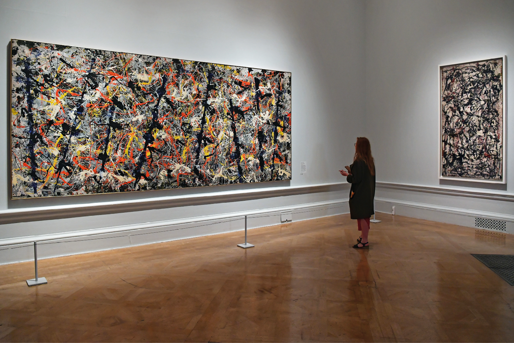 Fotografia. Uma mulher usando roupa preta está em um museu e de frente para um quadro retangular com uma pintura abstrata feita com a técnica do gotejamento de tinta. À sua direita, há outro quadro com o mesmo estilo.