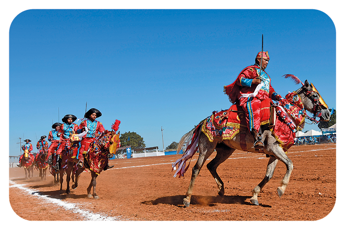 Fotografia. À direita, destaque para um homem usando fantasia azul e vermelha sobre cavalo com tecidos em tons de vermelho. À esquerda uma fileira de homens montados em cavalos e vestindo o mesmo traje.