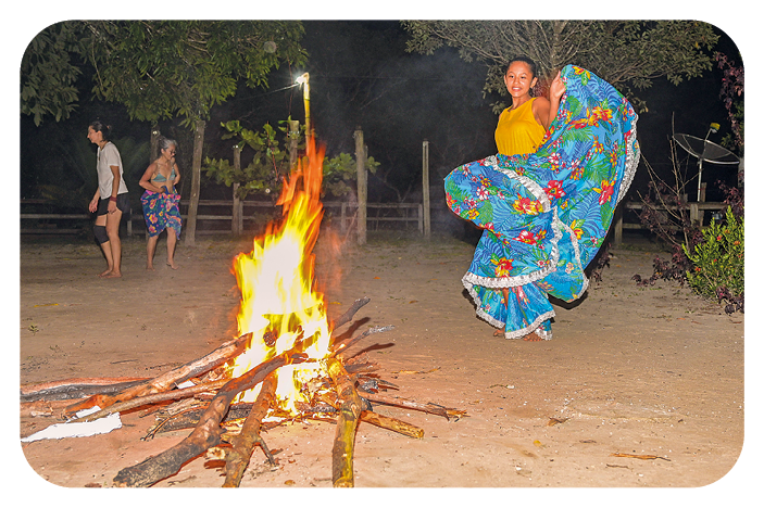 Fotografia. Uma mulher usando regata amarela e saia colorida em tons de azul está dançando com as mãos segurando a barra da saia na frente de uma fogueira. Ao fundo, há uma cerca de madeira e uma vegetação verde.
