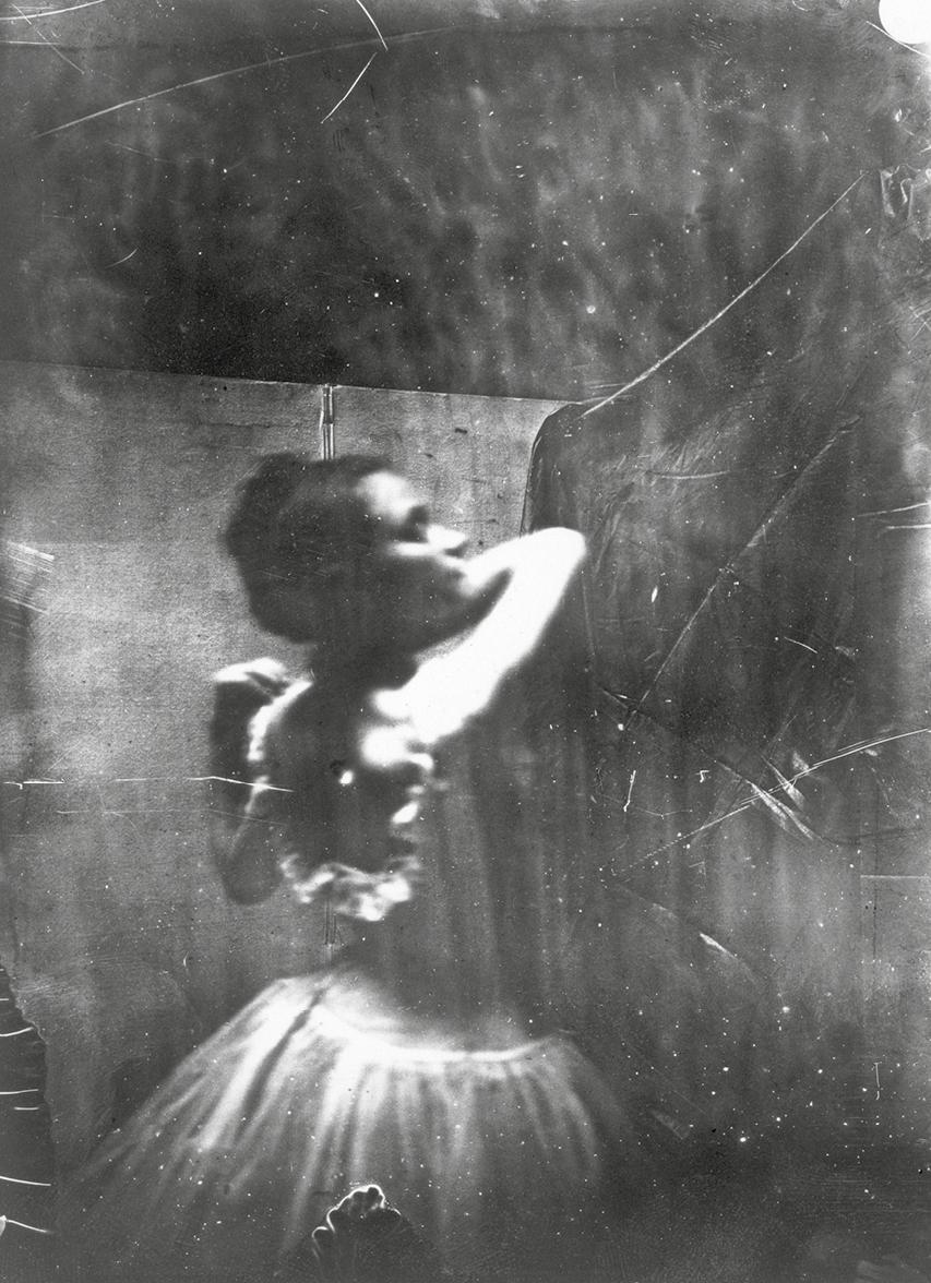 Fotografia em preto e branco. Vista de costas de uma mulher de cabelo preto penteado para trás, usando vestido branco, está com os dois braços flexionados para trás das costas e o rosto inclinado para a direita. Ao redor há linhas e manchas escuras.