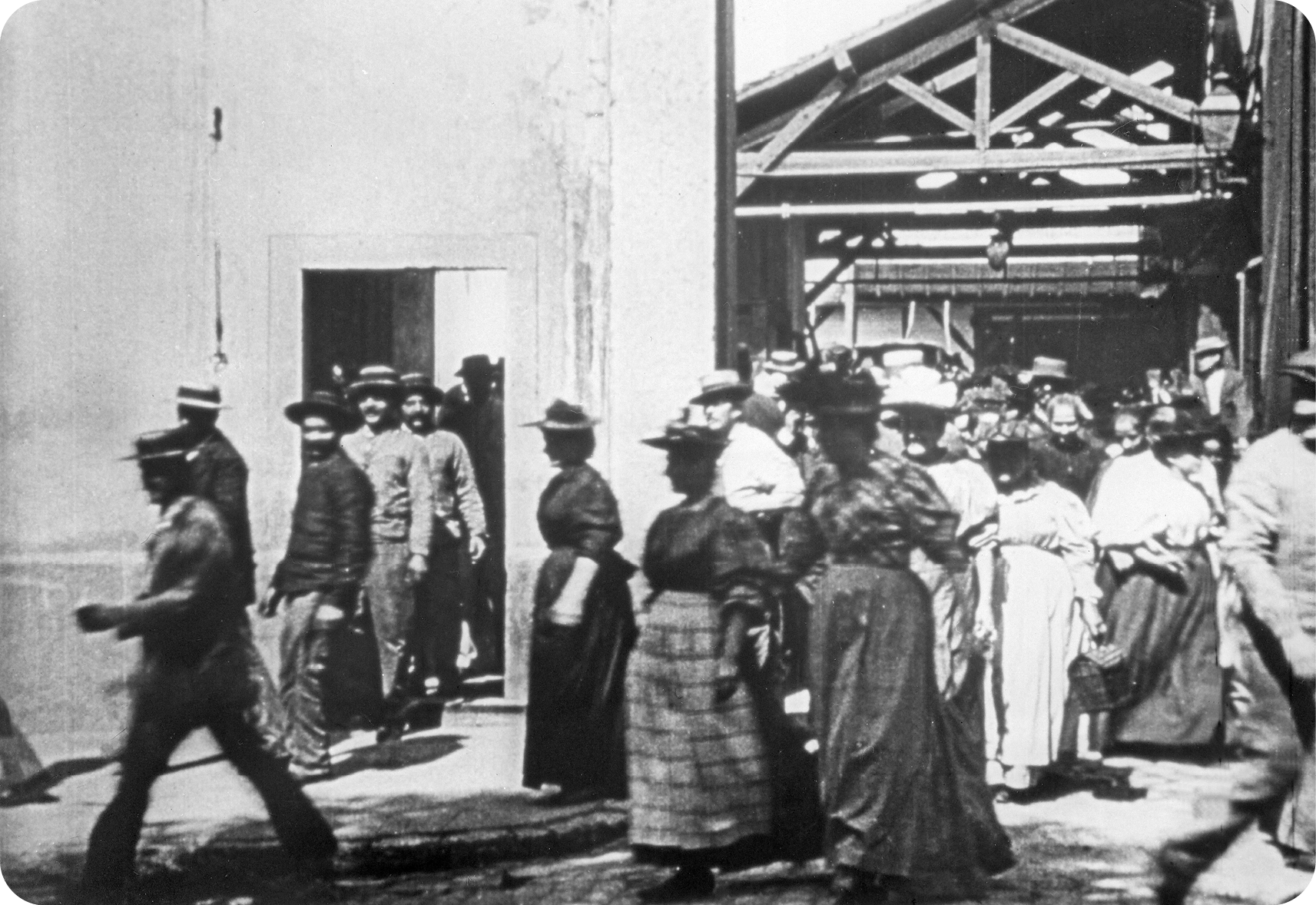 Fotografia em preto e branco. Vista de uma multidão de pessoas saindo por portas e galpões. Os homens usam chapéus redondos, camisa e calça; as mulheres usam chapéus finos, vestidos e saias. Ao fundo há uma grande construção de madeira.