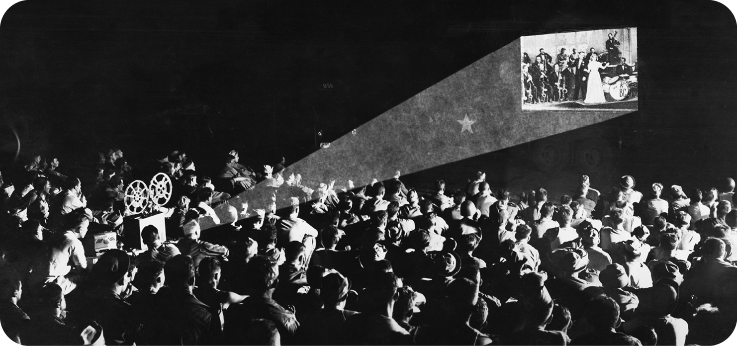 Fotografia em preto e branco. Vista de cima de uma multidão de pessoas sentadas; à esquerda, há um projetor quadrado com duas engrenagens redondas que produzem um feixe de luz em uma parede e projeta um filme.