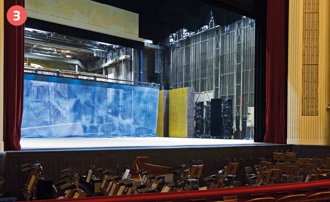 Fotografia. Vista parcial de um teatro. Em destaque, as cadeiras marrons de uma plateia e, ao fundo, um palco grande com cortinas vermelhas abertas nas laterais e um ciclorama ao fundo.