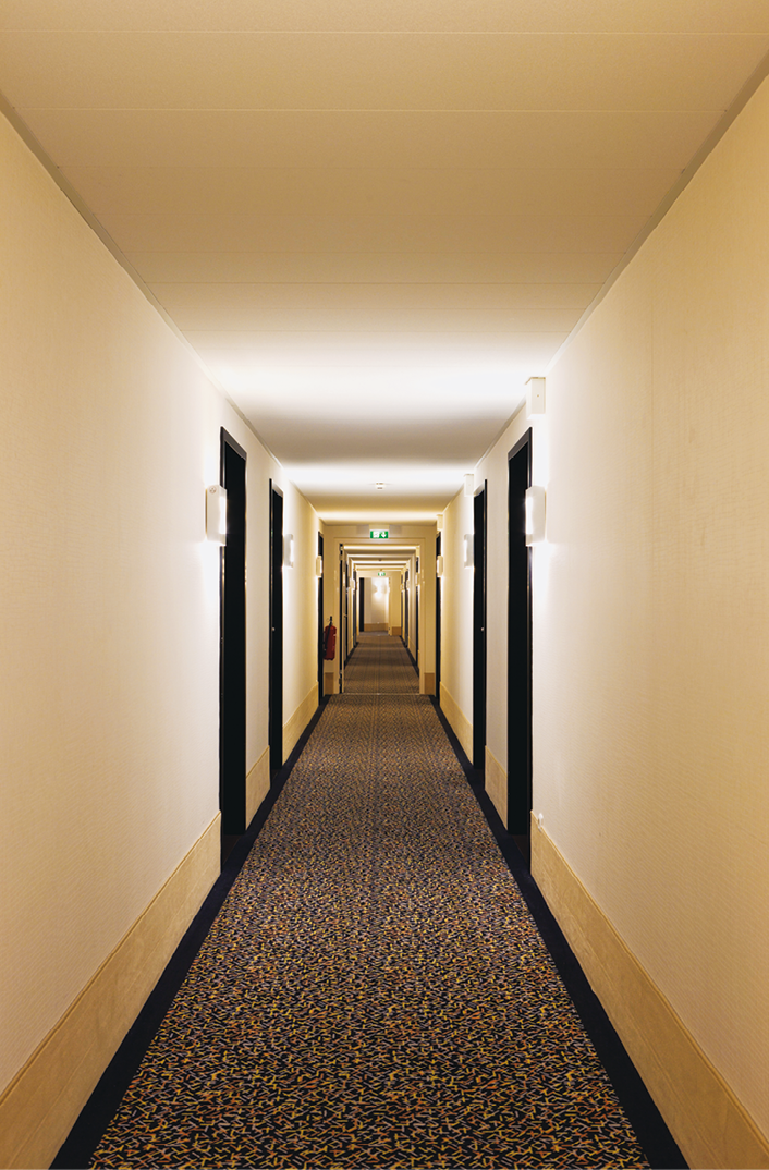 Fotografia. Vista em perspectiva de um corredor com paredes amareladas e portas pretas; conforme o corredor se distancia ao fundo, ele fica menor.