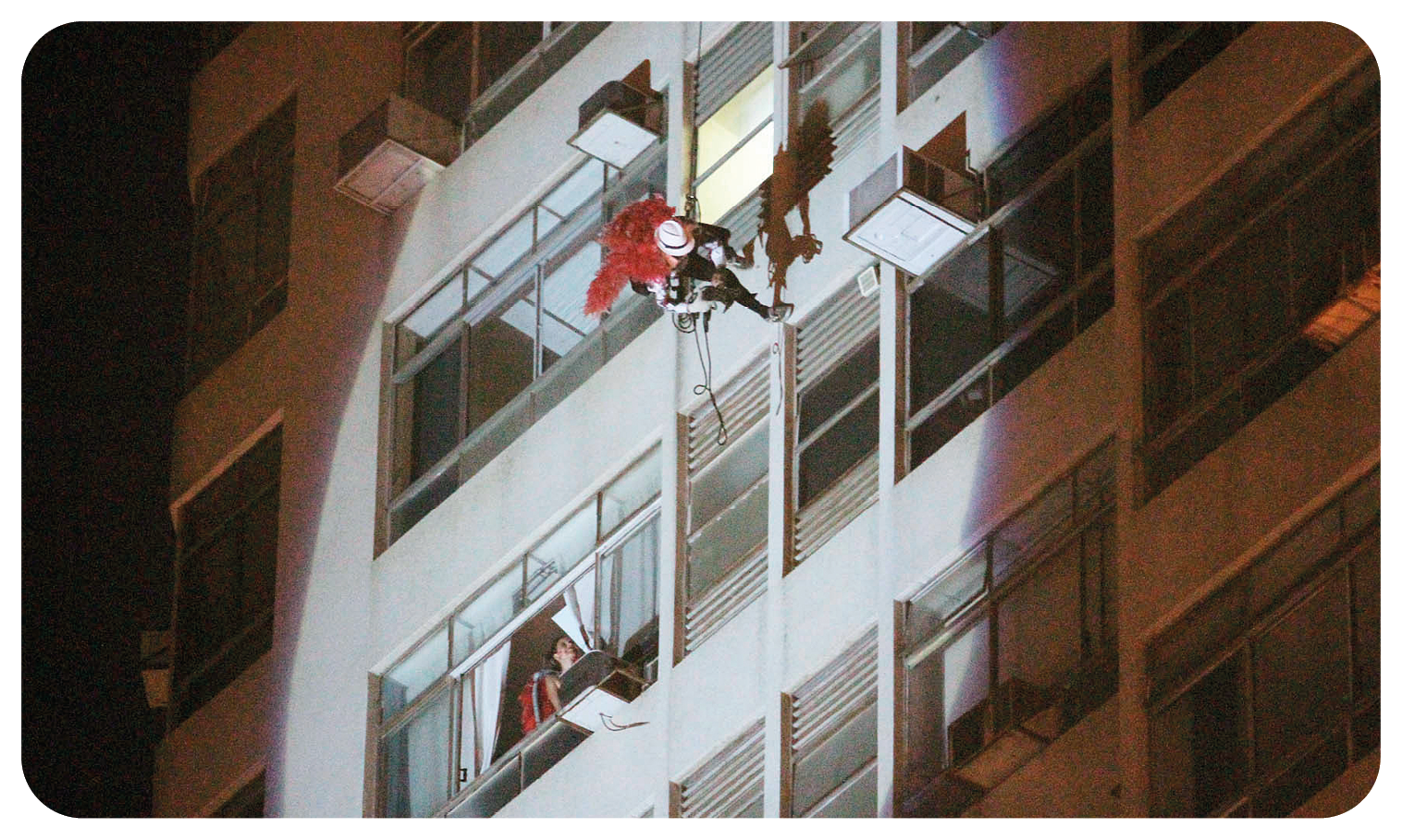 Fotografia. Vista de baixo para cima de um homem usando chapéu branco, roupa preta e capa vermelha; ele está descendo um prédio usando uma corda. À esquerda, há uma janela aberta onde uma mulher olha para o homem descendo. A cena é noturna; o homem e a mulher estão iluminados por uma luz que vem de baixo para cima.