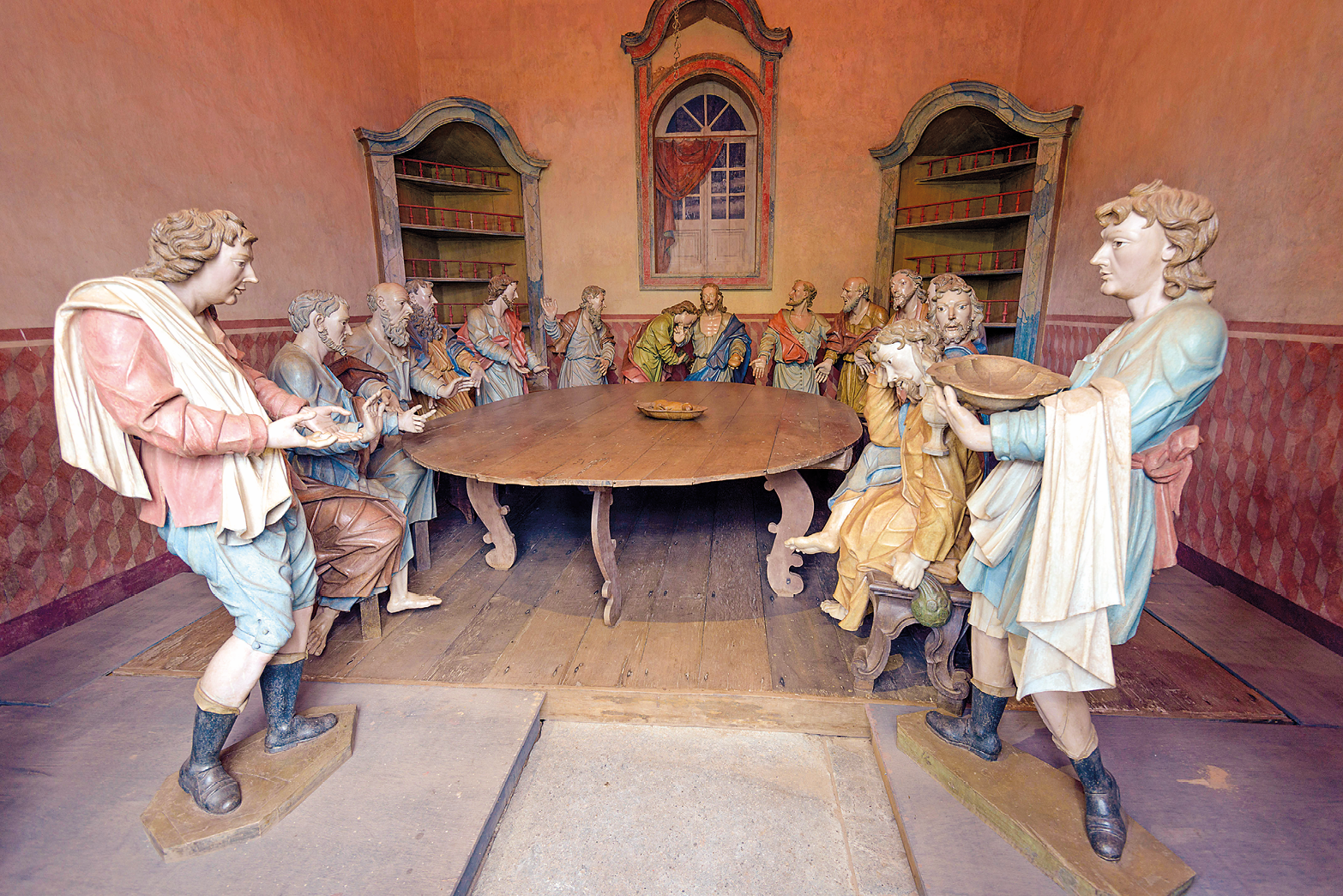 Fotografia. Escultura entalhada na madeira retratando um grupo de homens em pé ao redor de uma mesa redonda de madeira marrom. Eles vestem roupas coloridas e a maioria está com as mãos estendidas na direção da mesa