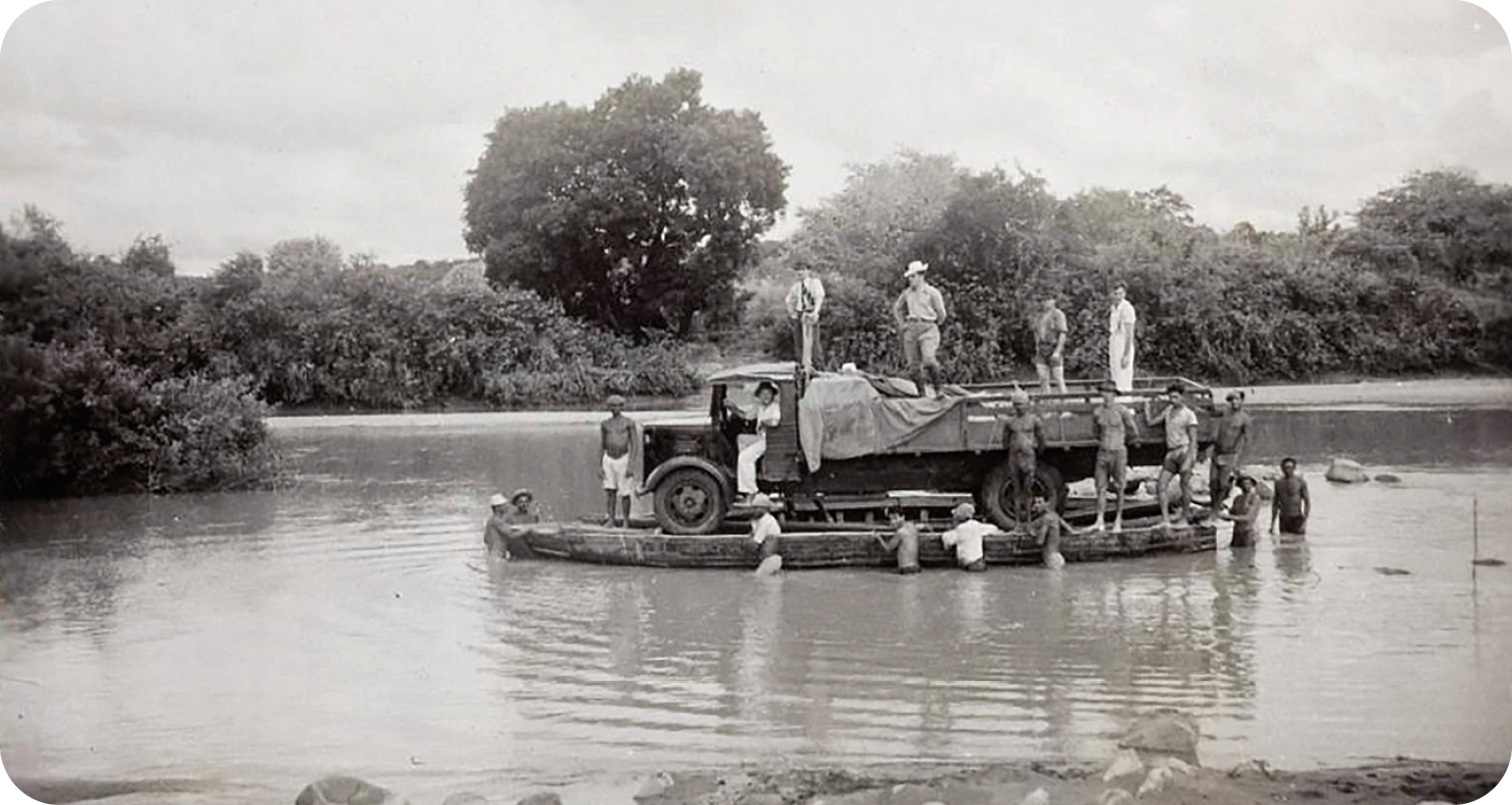 Fotografia em preto e branco. Vista de um rio com um caminhão sobre uma balsa e diversas pessoas em pé na balsa ou dentro do caminhão; algumas estão dentro do rio e empurram a balsa para a margem. Ao fundo, diversas árvores.