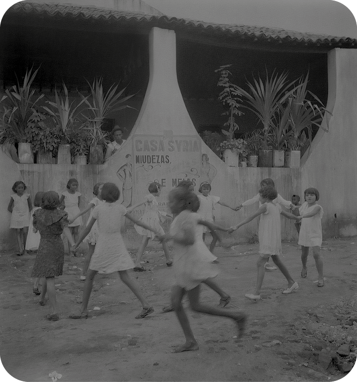 Fotografia em preto e branco. Destaque para diversas crianças - a maioria delas usando vestidos claros - e de mãos dadas formando uma roda.