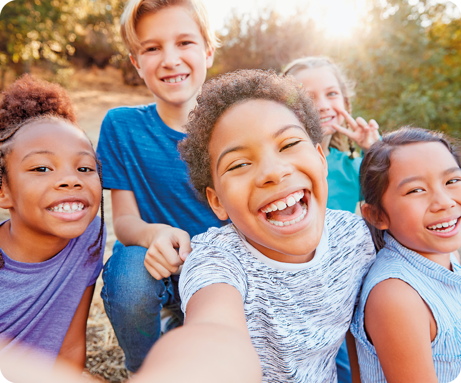 Fotografia. Destaque para cinco crianças com roupas coloridas. A que está no centro da imagem tira selfie enquanto todas olham para a câmera e sorriem. Ao fundo há um terreno com vegetação verde.