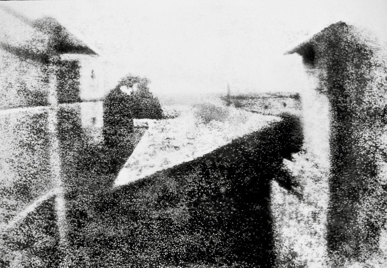 Fotografia em preto e branco. Vista do alto de uma rua. Nas laterais há prédios. Na parte inferior central, sombra.