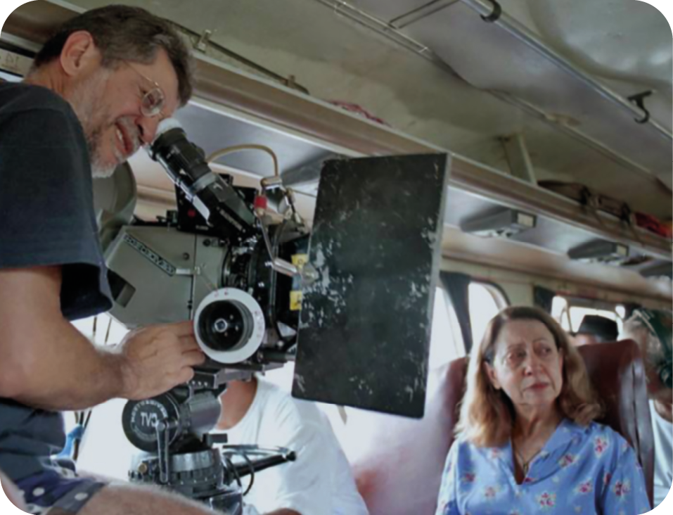 Fotografia. Making off da gravação de um filme. Um homem de cabelo grisalho, usando camiseta verde, está com a cabeça inclinada na direção de uma filmadora grande; filmando uma atriz de cabelo castanho e sentada em uma poltrona de um ônibus.