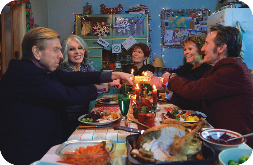 Cena de filme. No centro, uma mesa de jantar retangular com diversos pratos, copos, comidas e velas acesas. Ao redor da mesa, há três mulheres e dois homens sorrindo.