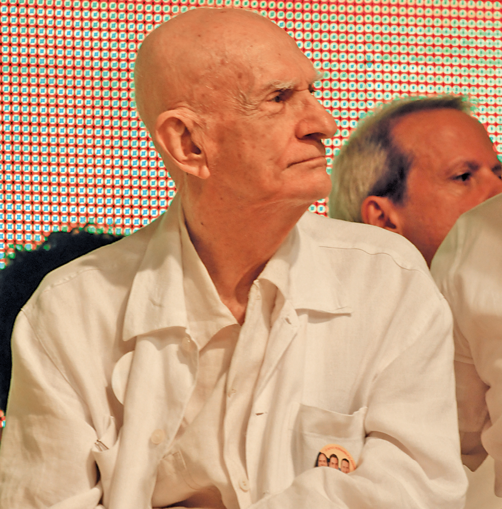 Fotografia. Um homem sem cabelos, de sobrancelha grossa e branca, nariz comprido, lábios finos, usando camisa branca, está com o cenho contraído e a cabeça inclinada para o lado.
