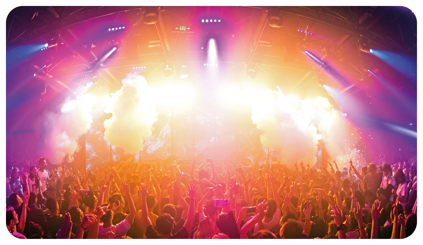Fotografia. Vista panorâmica de uma multidão de pessoas em pé com os braços levantados; na frente há um palco com diversas luzes e fumaças coloridas em tons de laranja, roxo, rosa e azul.