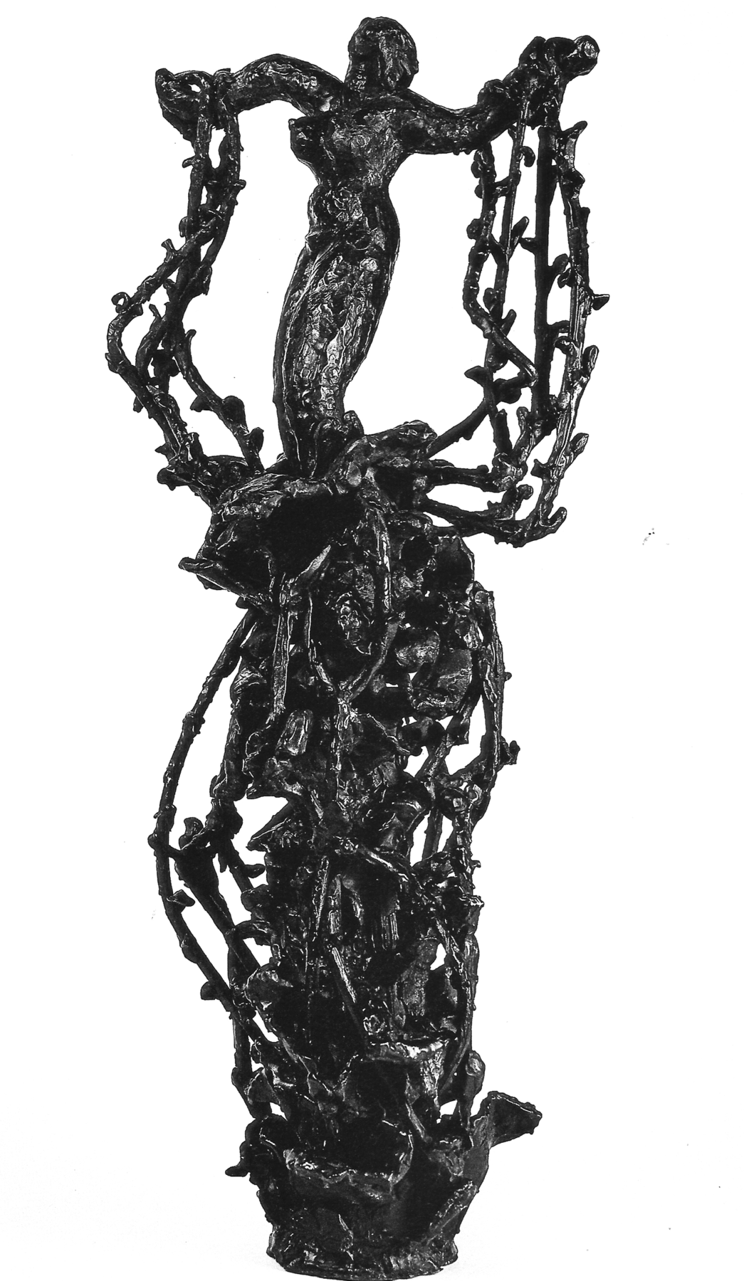 Fotografia. Escultura escura que apresenta uma mulher em pé sobre uma estrutura alta com muitos ramos entrelaçados. Ela está com os braços abertos para o lado e com os pulsos presos pelos ramos.
