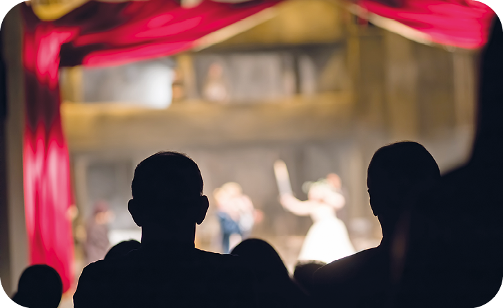 Fotografia. Vista do fundo para a frente de uma plateia no escuro que observa uma peça no palco ao fundo, emoldurado por uma cortina vermelha.