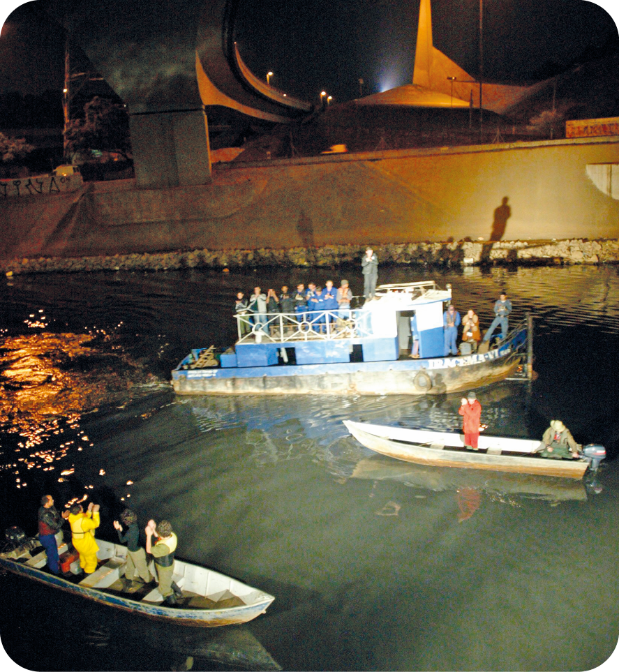 Encenação. A cena é apresentada durante a noite em um rio, onde há três embarcações de tamanhos diferentes e com pouca tripulação. O local perto dos barcos está iluminado. Ao fundo, está uma parede de concreto..