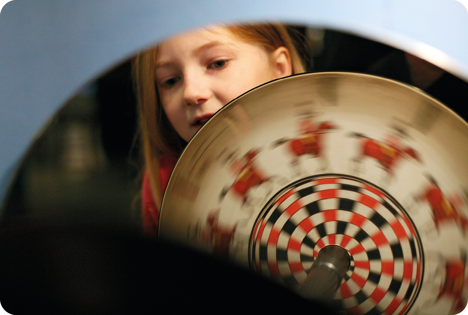 Fotografia. Destaque de um espelho mostrando a imagem de uma menina segurando um objeto circular com desenhos semelhantes em sua superfície, o qual está em movimento.