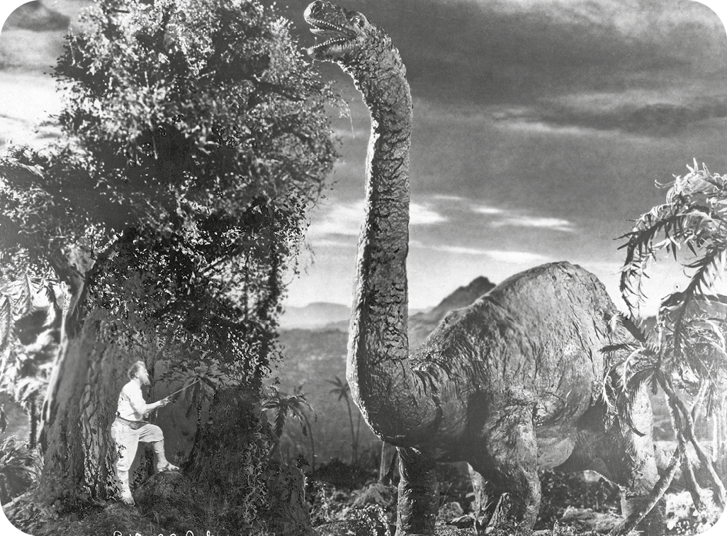 Cena de filme em preto e branco. À direita, um dinossauro de corpo robusto e longo pescoço se alimenta das folhas de uma árvore. À esquerda, uma pessoa em tamanho bem menor e vestindo roupas claras observa parada ao lado do tronco de uma árvore.