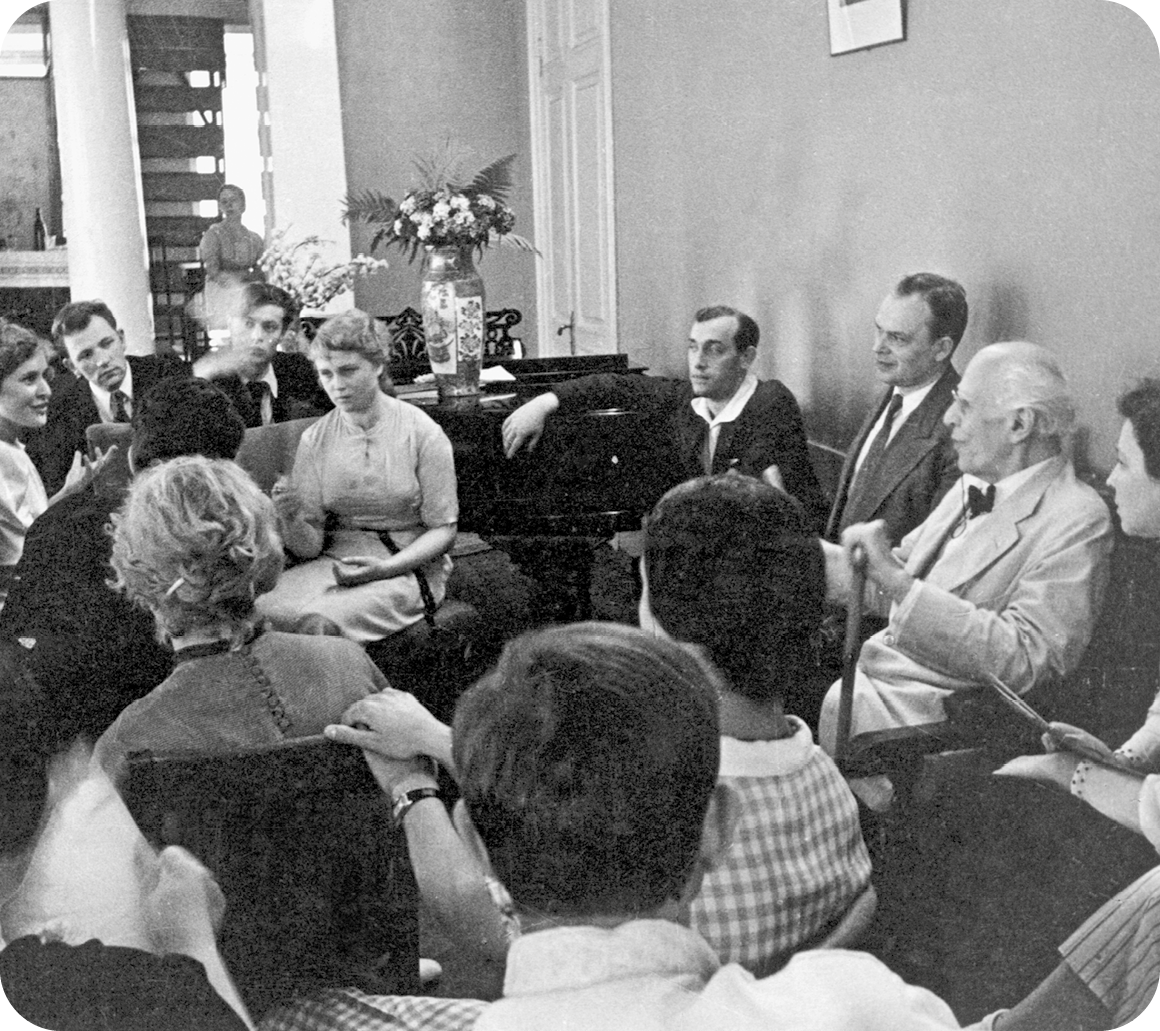 Fotografia em preto e branco. No interior de uma sala, destaque para um senhor de cabelos grisalhos e roupas claras segurando uma bengala e sentado à direita. Ao redor dele, há vários homens e mulheres sentados e conversando.