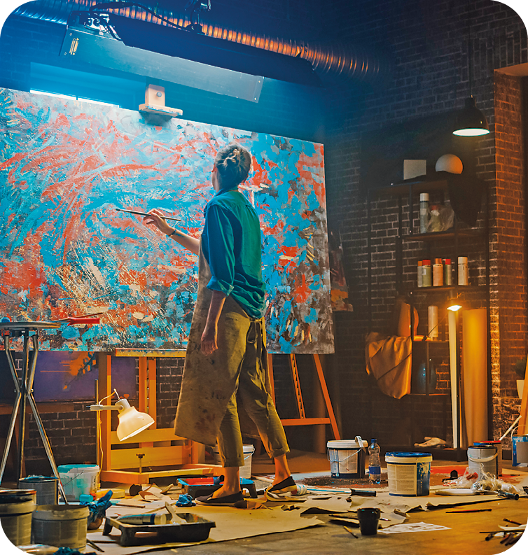 Fotografia. Em uma sala fechada com luz baixa, uma pessoa está em pé e pinta uma ampla obra abstrata nas cores azul e laranja. No chão, há baldes de tinta e pincéis.