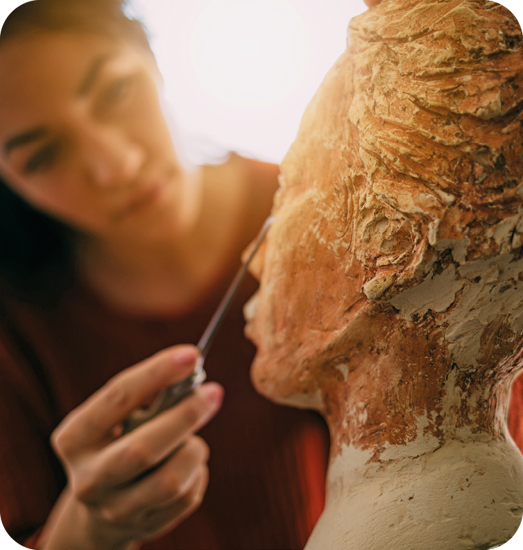 Fotografia. Destaque da mão de uma mulher realizando os detalhes em uma escultura que apresenta um rosto humano.