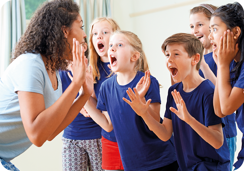 Fotografia. À esquerda, uma mulher de cabelos encaracolados, vista de lado, está com a boca aberta e as mãos espalmadas diante de um pequeno grupo de crianças que imitam sua expressão; elas estão uniformizadas com blusa azul.